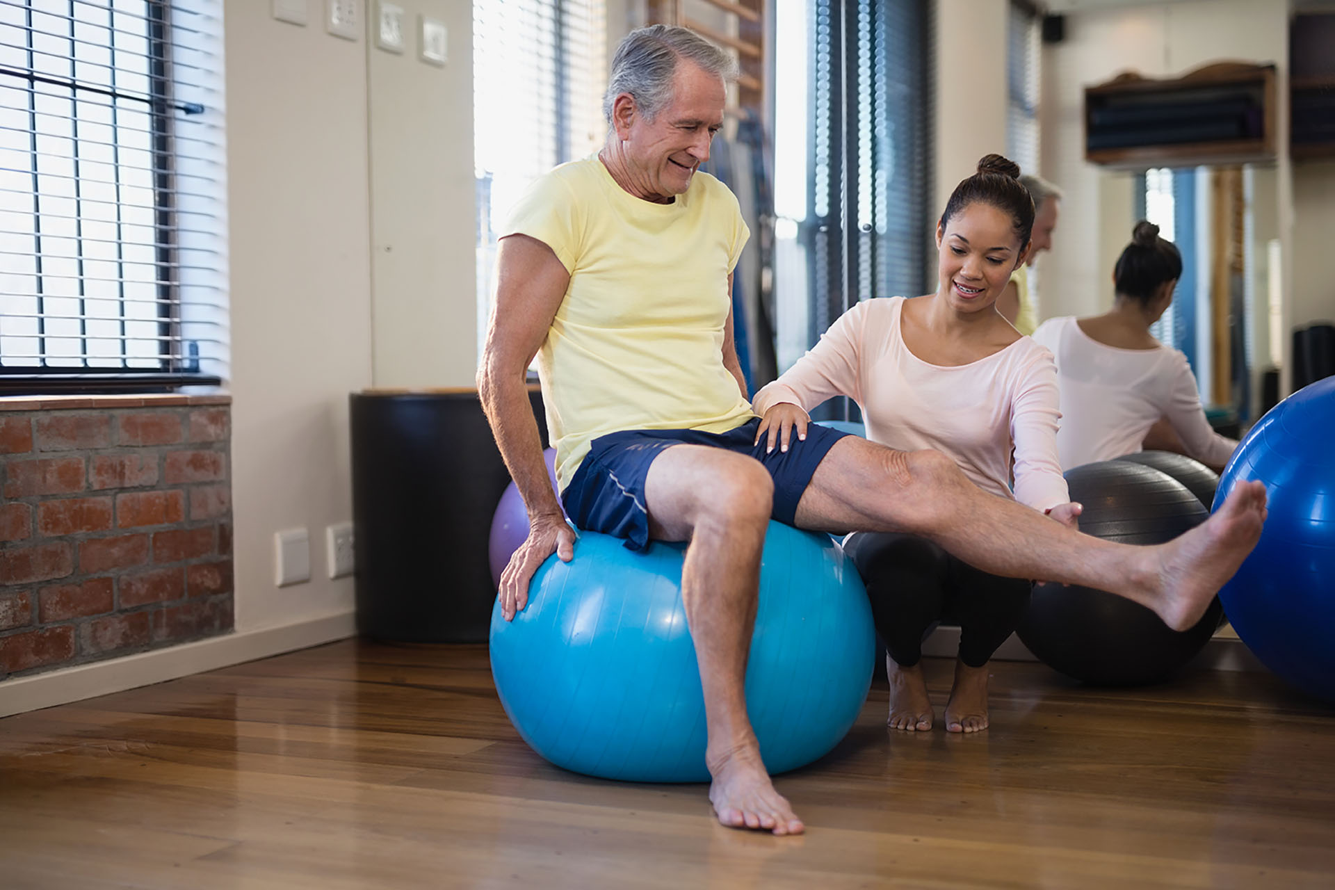 Female therapist helping senior male patient doing leg exercise on blue ball